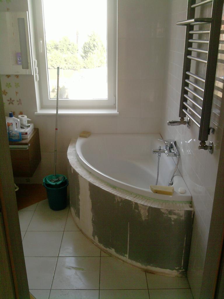 Narożna wanna przed wykończeniem w łazience z oknem. Widoczne ślady szpachlowania na obudowie wanny i mop w wiadrze. Grzejnik drabinkowy na ścianie.