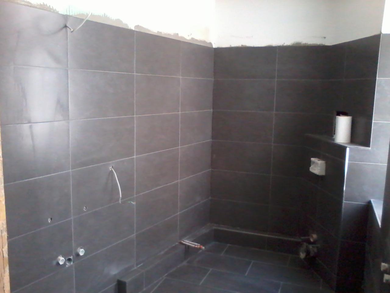 Ciemnoszara, wykafelkowana łazienka w trakcie remontu. Widoczne niedokończone fugi, wystające kable i rury. Na półce rolka papieru toaletowego.