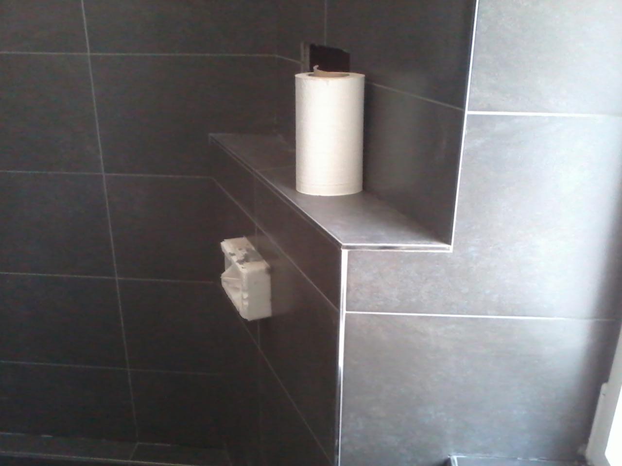 Półka w łazience wykończona szarymi płytkami z rolką papieru i dozownikiem. Widoczne fugi i kąt ujęcia podkreślają detale wykończenia wnętrza.