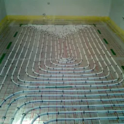 Białe rury ogrzewania podłogowego ułożone spiralnie na folii z siatką, zabezpieczone taśmą przy ścianach. Widok z góry na instalację przed wylaniem posadzki.