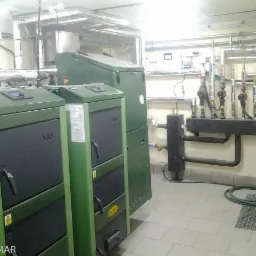 Zielone kotły grzewcze w kotłowni z rurami i osprzętem. Widoczne elementy instalacji grzewczej na ścianie. Kotły marki Brosol z czarnymi drzwiczkami.