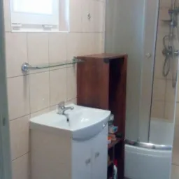 Jasna łazienka z umywalką na szafce, kabiną prysznicową i brązowym regałem. Beżowe płytki na ścianach i dywanik na podłodze. Widoczne okno.