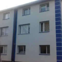 Ocieplony budynek wielorodzinny z nową elewacją w odcieniach bieli i błękitu. Widoczne okna i balkon. Drzewa w tle.