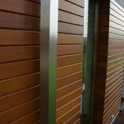 Elewacja budynku wykończona poziomymi deskami w ciepłym odcieniu brązu, z widoczną aluminiową kolumną i fragmentem okna. Deski tworzą regularny wzór, dodając elegancji.