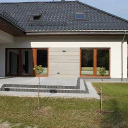 Nowoczesny dom jednorodzinny z szarym dachem, drewnianą stolarką okienną i brukowanym tarasem, otoczony trawnikiem z młodymi drzewkami. Widok z zewnątrz.