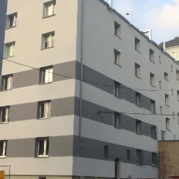 Elewacja budynku wielorodzinnego w Ćwiklicach po tynkowaniu, z poziomymi pasami szarości i bieli. Widoczne rusztowania z lewej strony budynku.