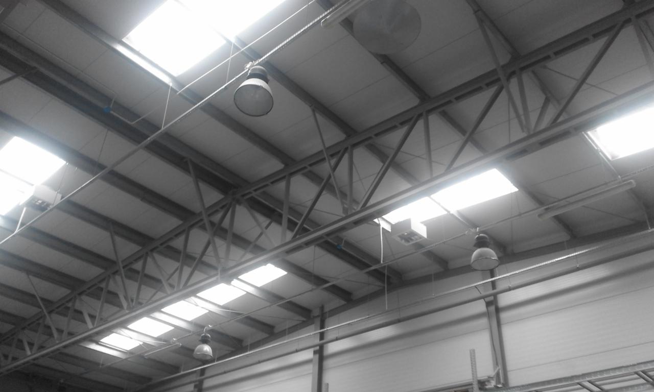 Wnętrze hali magazynowej z widoczną konstrukcją dachu, oświetleniem przemysłowym i świetlikami dachowymi.