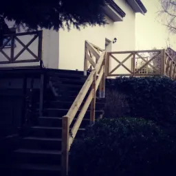 Drewniane schody z balustradą w stylu rustykalnym prowadzą do domu. Balustrada z krzyżowych elementów. Oświetlenie zewnętrzne. Zieleń w tle.