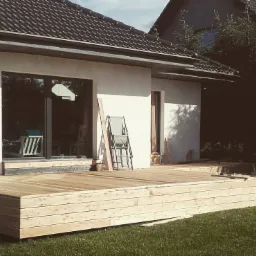 Jasny taras z desek przy białym domu z ciemną dachówką, z krzesłem i deskami opartymi o ścianę. Widok na trawnik. Ukończony etap budowy.