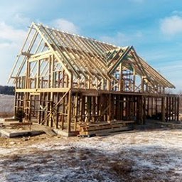 Marcin Pląskowski - Szkielet domu w trakcie budowy, widoczne drewniane elementy konstrukcyjne na fundamencie, zimowy krajobraz w tle, błękitne niebo. Dom w stylu nowoczesnym.