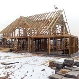 Marcin Pląskowski - Surowy szkielet drewniany domu w budowie, widoczne krokwie dachu i elementy konstrukcyjne. Na pierwszym planie ułożone płyty chodnikowe, w tle krajobraz.