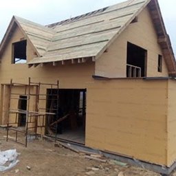 Marcin Pląskowski - Dwu kondygnacyjny dom szkieletowy w trakcie budowy. Widoczne elementy konstrukcyjne ścian i dachu. Surowy stan, brak okien i drzwi. Teren budowy wokół.