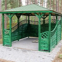Zielona altana ogrodowa z drewnianą konstrukcją i ażurowymi panelami, otoczona drzewami. Ścieżka z kostki brukowej prowadzi do wejścia.