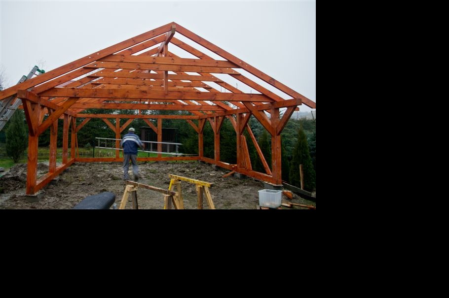 Konstrukcja drewniana altany w trakcie budowy, widoczne elementy szkieletu i pracownik z drabiną. Na pierwszym planie narzędzia i teren budowy.