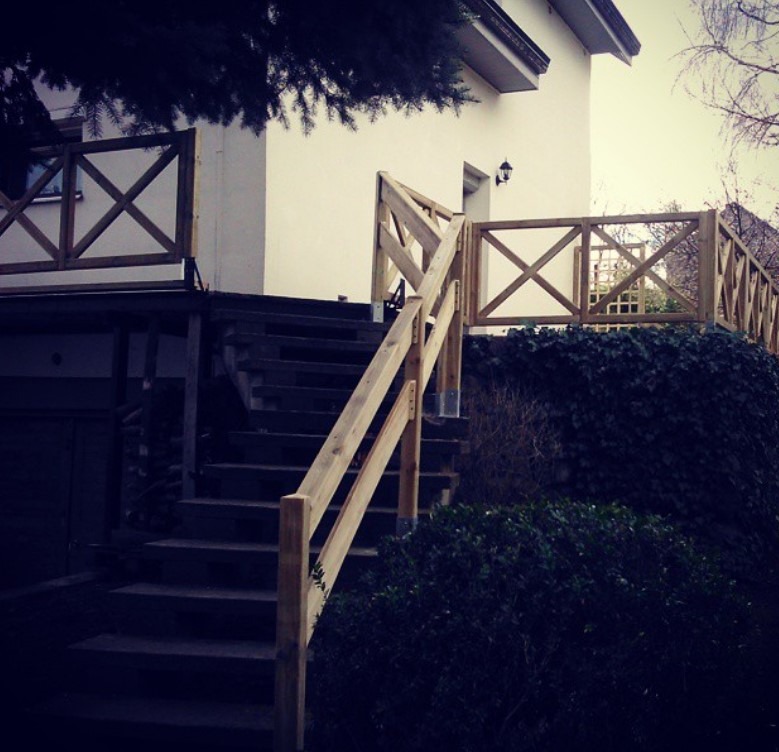 Drewniane schody z balustradą w stylu rustykalnym prowadzą do domu. Balustrada z krzyżowych elementów. Oświetlenie zewnętrzne. Zieleń w tle.