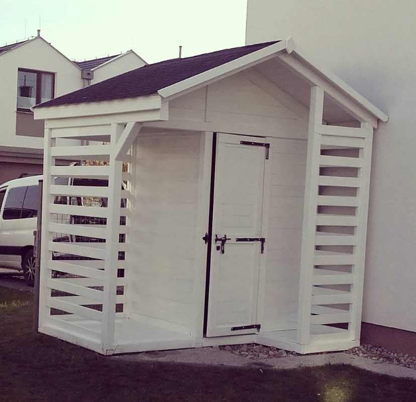 Biała, drewniana konstrukcja z drzwiami i ażurowymi ścianami bocznymi, przypominająca mały domek ogrodowy na tle budynku mieszkalnego i zieleni.