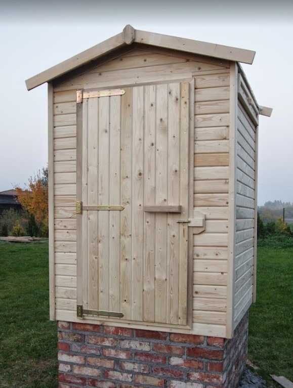 Drewniana budka narzędziowa na fundamencie z cegieł, z metalowymi zawiasami, w Starogardzie Gdańskim. Solidna konstrukcja, widoczne usłojenie drewna.