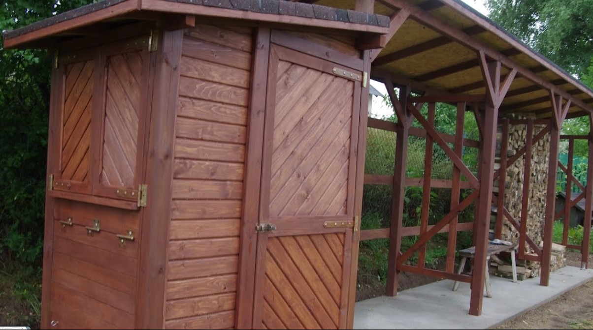 Drewniana altana z zamykaną częścią, brązowa impregnacja, widoczne detale okuć i konstrukcji dachu, na betonowej wylewce, z zapasem drewna w tle.