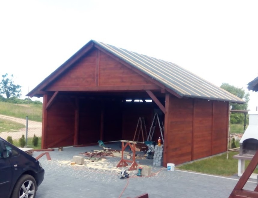 Ciemnobrązowa drewniana konstrukcja z dachem w trakcie budowy, widoczne narzędzia i materiały budowlane, teren posesji, częściowo widoczny samochód.