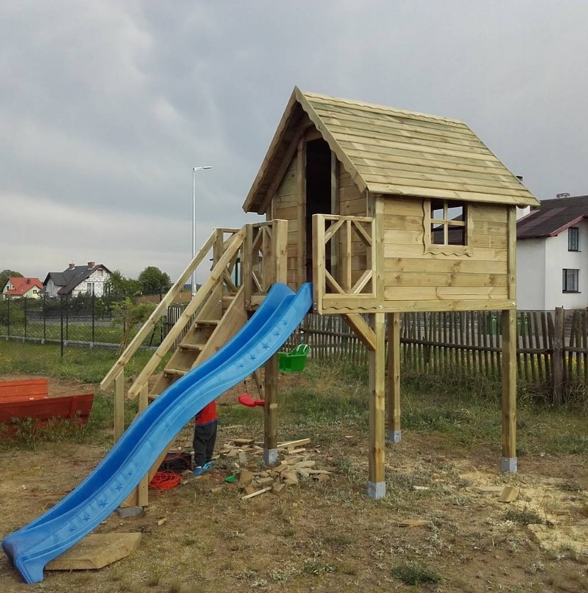 Drewniany domek dla dzieci na podwyższeniu z niebieską zjeżdżalnią, schodami i balustradą, na tle szarego nieba i zabudowań. Widoczne detale wykończenia i częściowo osoba.