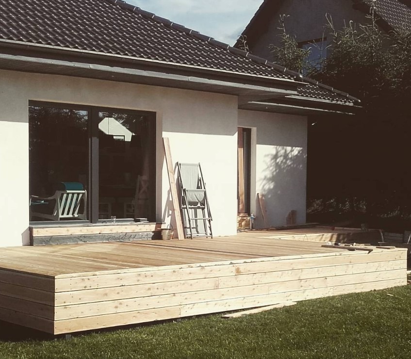 Jasny taras z desek przy białym domu z ciemną dachówką, z krzesłem i deskami opartymi o ścianę. Widok na trawnik. Ukończony etap budowy.