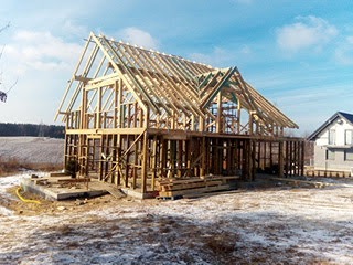 Szkielet domu w trakcie budowy, widoczne drewniane elementy konstrukcyjne na fundamencie, zimowy krajobraz w tle, błękitne niebo. Dom w stylu nowoczesnym.