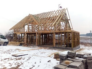 Surowy szkielet drewniany domu w budowie, widoczne krokwie dachu i elementy konstrukcyjne. Na pierwszym planie ułożone płyty chodnikowe, w tle krajobraz.