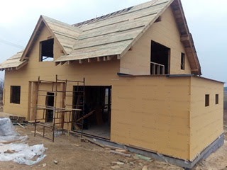 Dwu kondygnacyjny dom szkieletowy w trakcie budowy. Widoczne elementy konstrukcyjne ścian i dachu. Surowy stan, brak okien i drzwi. Teren budowy wokół.