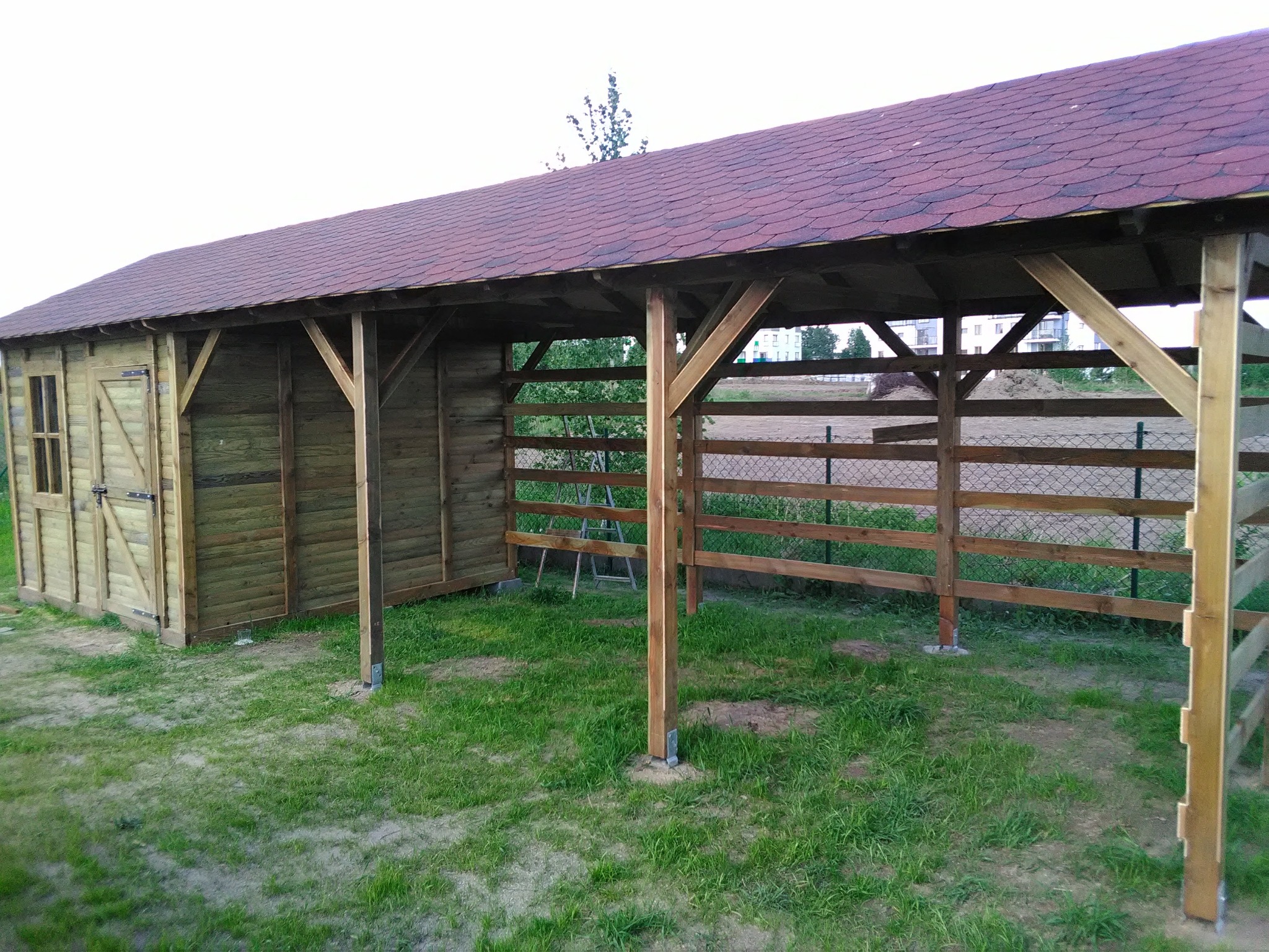 Drewniana konstrukcja z dachem z gontu bitumicznego, częściowo zabudowana, z widocznymi elementami więźby dachowej i słupami na zielonym trawniku.