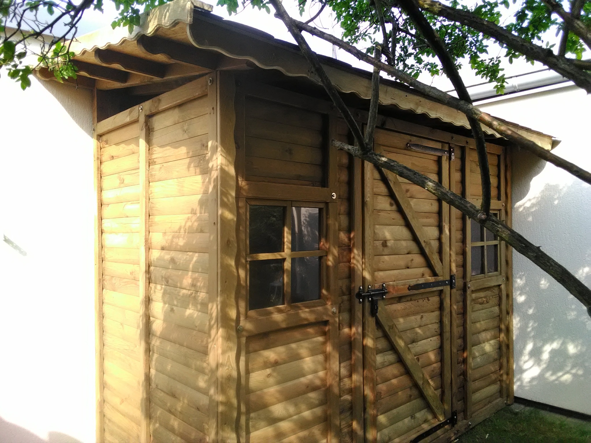 Drewniana altanka z oknem i dwuskrzydłowymi drzwiami, z dekoracyjnym wykończeniem dachu, na tle białej ściany i zieleni, częściowo przysłonięta gałęziami drzewa.