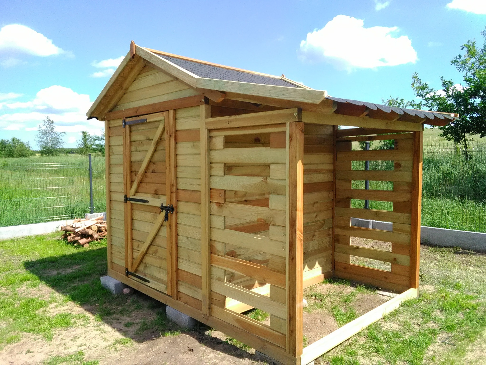 Drewniana konstrukcja z dachem krytym gontem bitumicznym, częściowo ażurowa, na trawniku. Widoczne drewniane elementy i zasuwa. W tle zielona trawa i błękitne niebo.