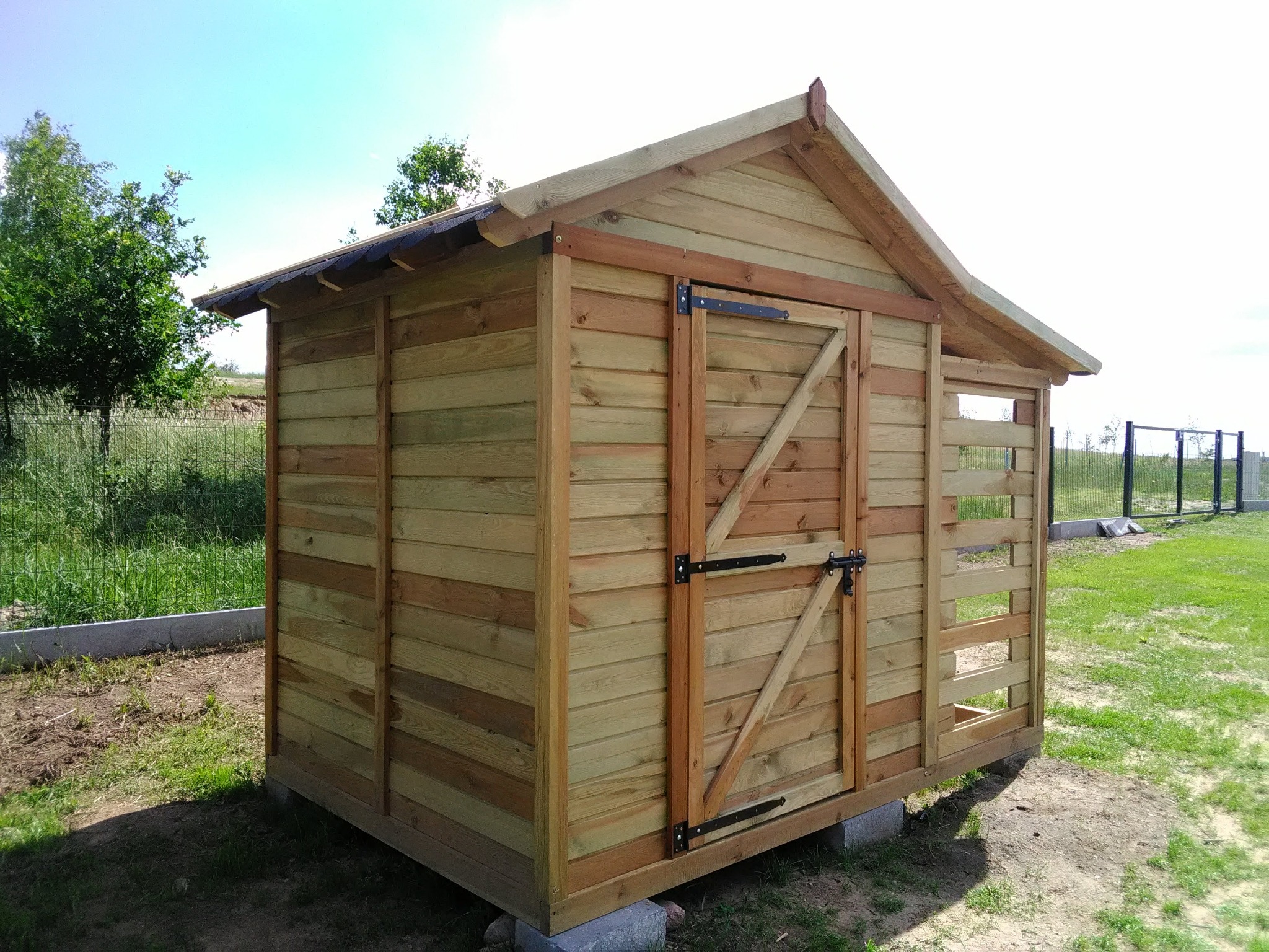 Drewniana konstrukcja z zamykanymi drzwiami, prawdopodobnie domek narzędziowy, wykonana z jasnego drewna, umieszczona na betonowych blokach na trawiastym terenie.