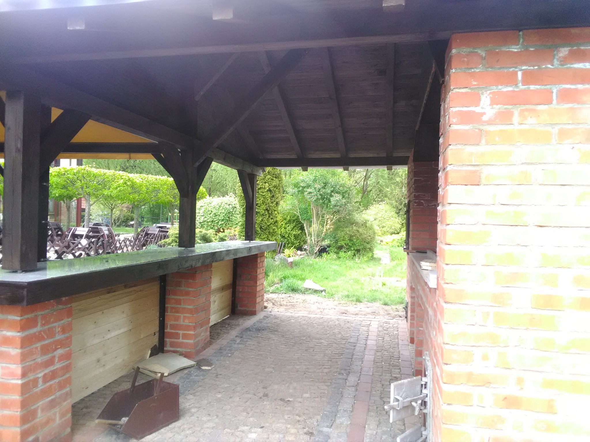 Ceglana konstrukcja z drewnianym zadaszeniem, widok na ogród. Fragment konstrukcji z blatem i drewnianym wypełnieniem. Ułożona kostka brukowa.
