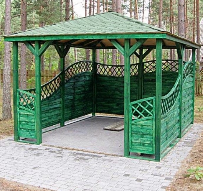 Zielona altana ogrodowa z drewnianą konstrukcją i ażurowymi panelami, otoczona drzewami. Ścieżka z kostki brukowej prowadzi do wejścia.