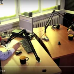 Studio nagrań z dwoma osobami przy biurkach, mikrofonami i słuchawkami, jedna osoba w trakcie mówienia do mikrofonu, druga siedzi naprzeciwko z zamazaną twarzą.