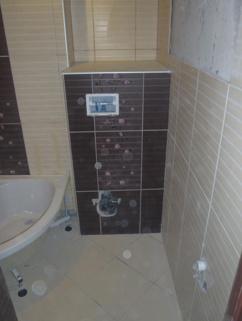 Wnętrze łazienki w trakcie prac wykończeniowych. Widoczne płytki w odcieniach beżu i brązu, częściowo zamontowana armatura oraz elementy instalacji sanitarnej.