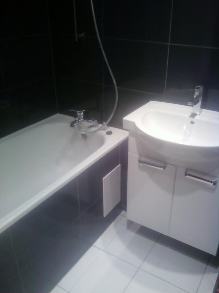 Biała wanna z baterią i umywalka z szafką w łazience wyłożonej czarnymi i białymi płytkami. Minimalistyczny design, chromowane elementy.