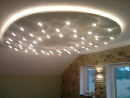 Oryginalny sufit z podwieszaną okrągłą konstrukcją i licznymi punktowymi lampami, ściana z cegły i dwa okna w pomieszczeniu.