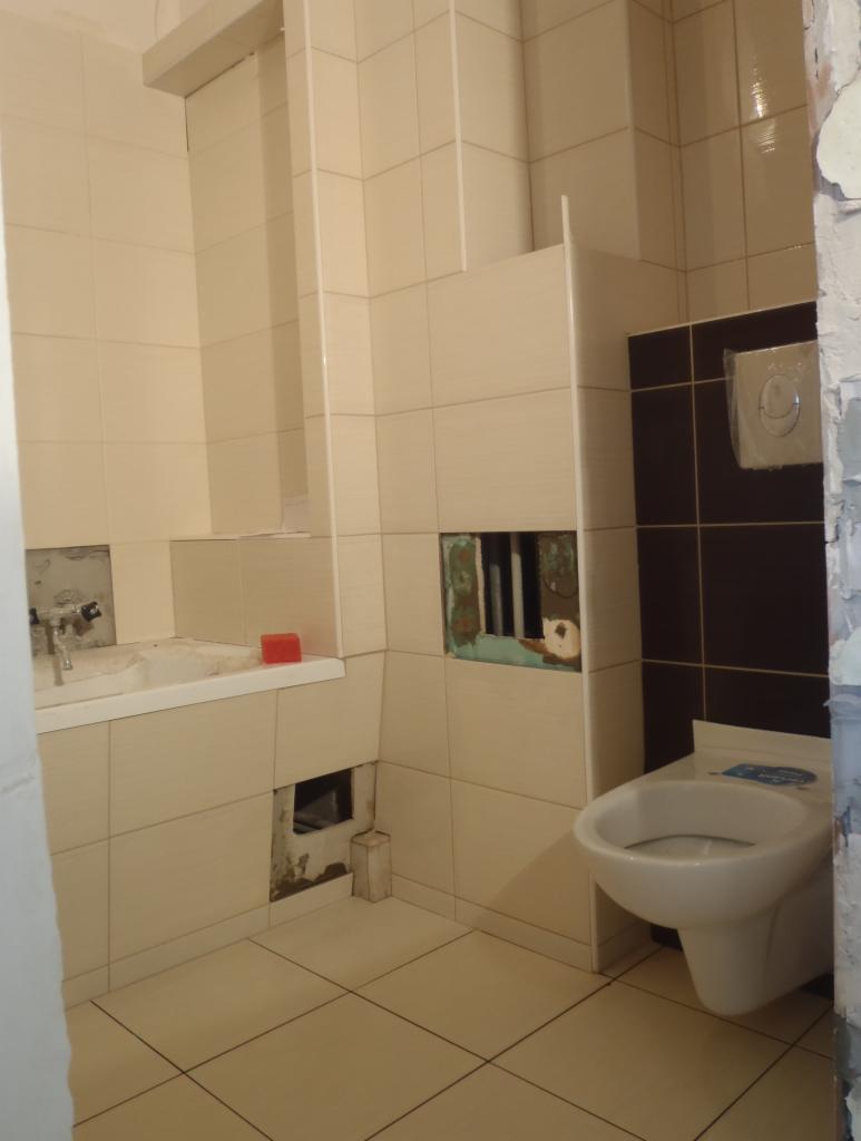 Łódź: Częściowo wykafelkowana łazienka w trakcie remontu. Widoczne niedokończone ściany, wanna i toaleta. Prace glazurnicze w toku, kremowe i brązowe płytki.