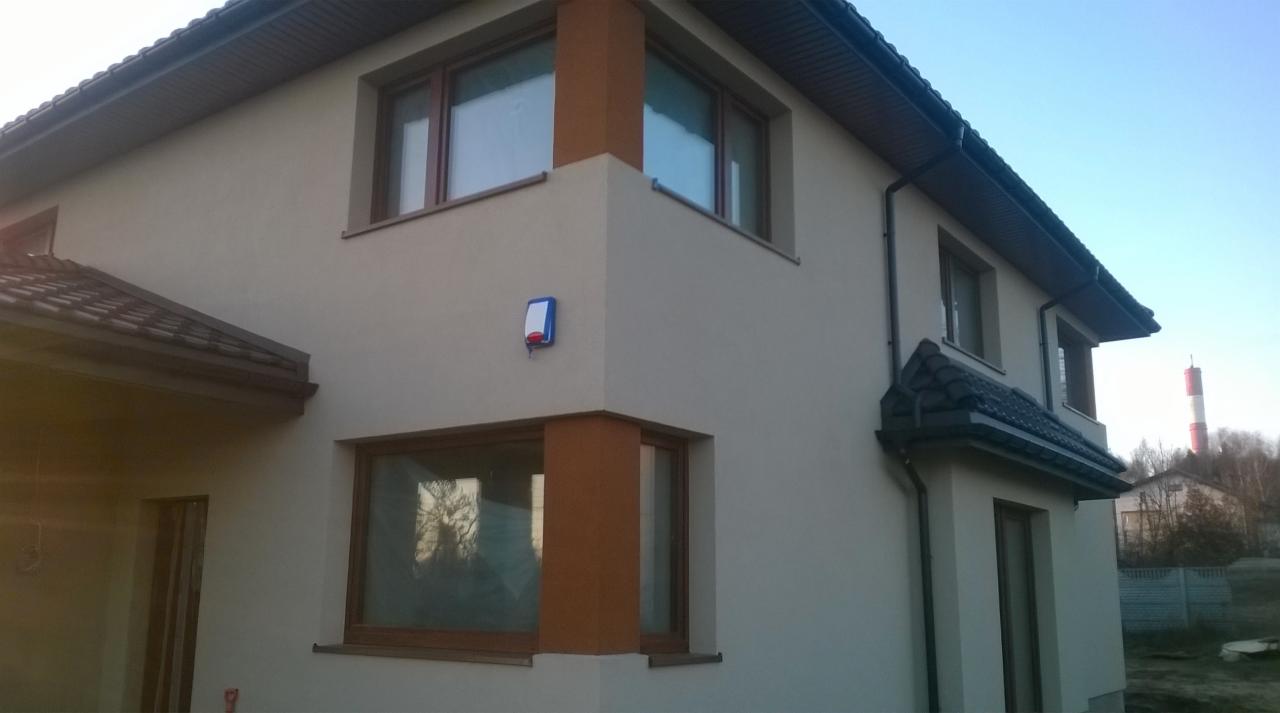 Dwukondygnacyjny dom z beżową elewacją, brązowymi ramami okiennymi i dachem z ciemnej dachówki. Widoczny fragment ogrodu i komin w tle.