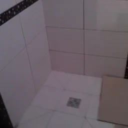 Wykończony brodzik z jasnymi płytkami i ozdobnym pasem z ciemnych płytek. Widoczny odpływ liniowy i fragment kartonu zabezpieczającego podłogę.