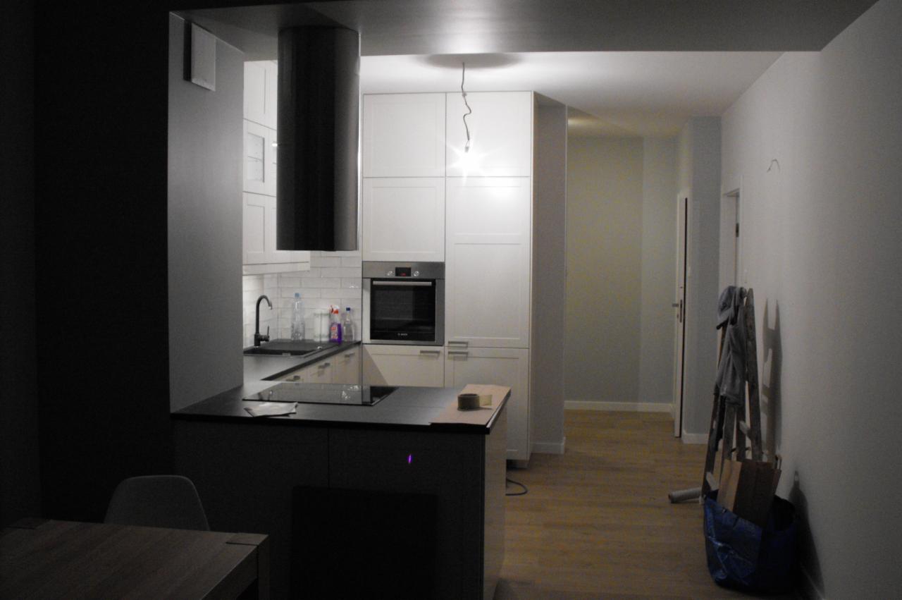 Nowoczesna kuchnia z białymi szafkami i czarnym okapem cylindrycznym. Widoczna wyspa kuchenna z blatem i fragment korytarza z drabiną.