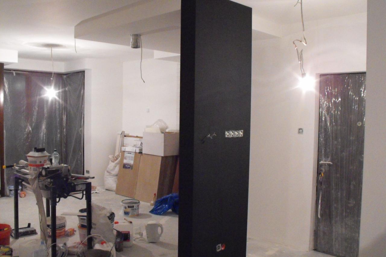Wnętrze w trakcie prac wykończeniowych: czarna kolumna, zabezpieczone folią drzwi, sprzęt malarski i kartony. Widoczne niedokończone instalacje elektryczne.