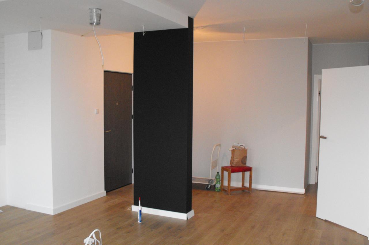 Wnętrze mieszkania w trakcie prac wykończeniowych: pomalowana na czarno ściana działowa, drewniana podłoga, widoczne narzędzia i materiały budowlane.