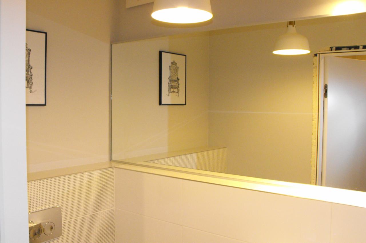 Nowoczesna łazienka z dużym lustrem, jasnymi płytkami i minimalistycznym oświetleniem. Na ścianie grafika w czarnej ramie. Widoczny fragment kabiny prysznicowej.