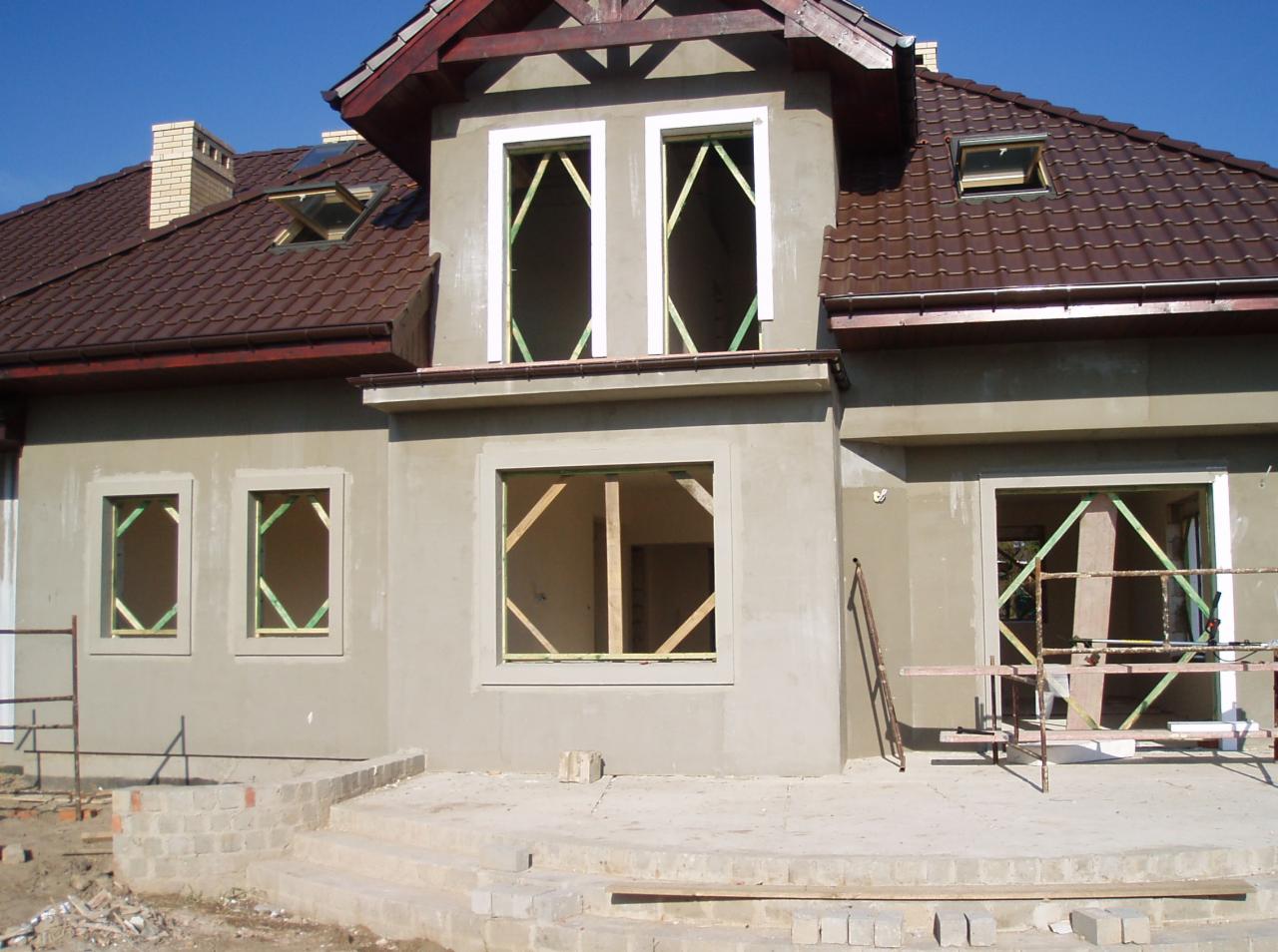 Dom w stanie surowym zamkniętym: szara elewacja, brązowy dach, widoczne drewniane ramy okienne. Częściowo otynkowane ściany, schody wejściowe w trakcie budowy.