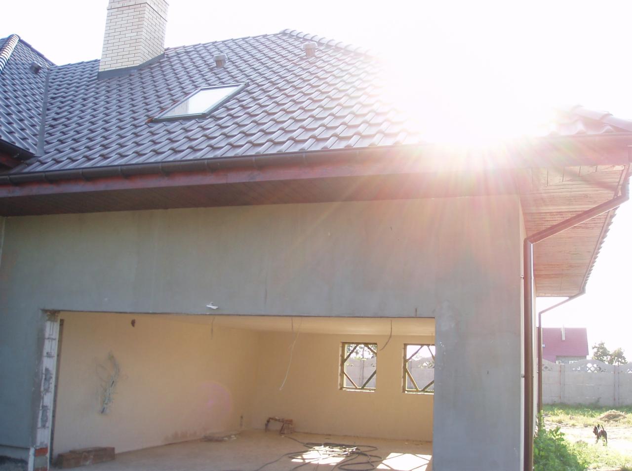 Surowy stan domu z ceglanym kominem i dachem z dachówki, widok na wnętrze przez otwór garażowy, wpadające promienie słoneczne.