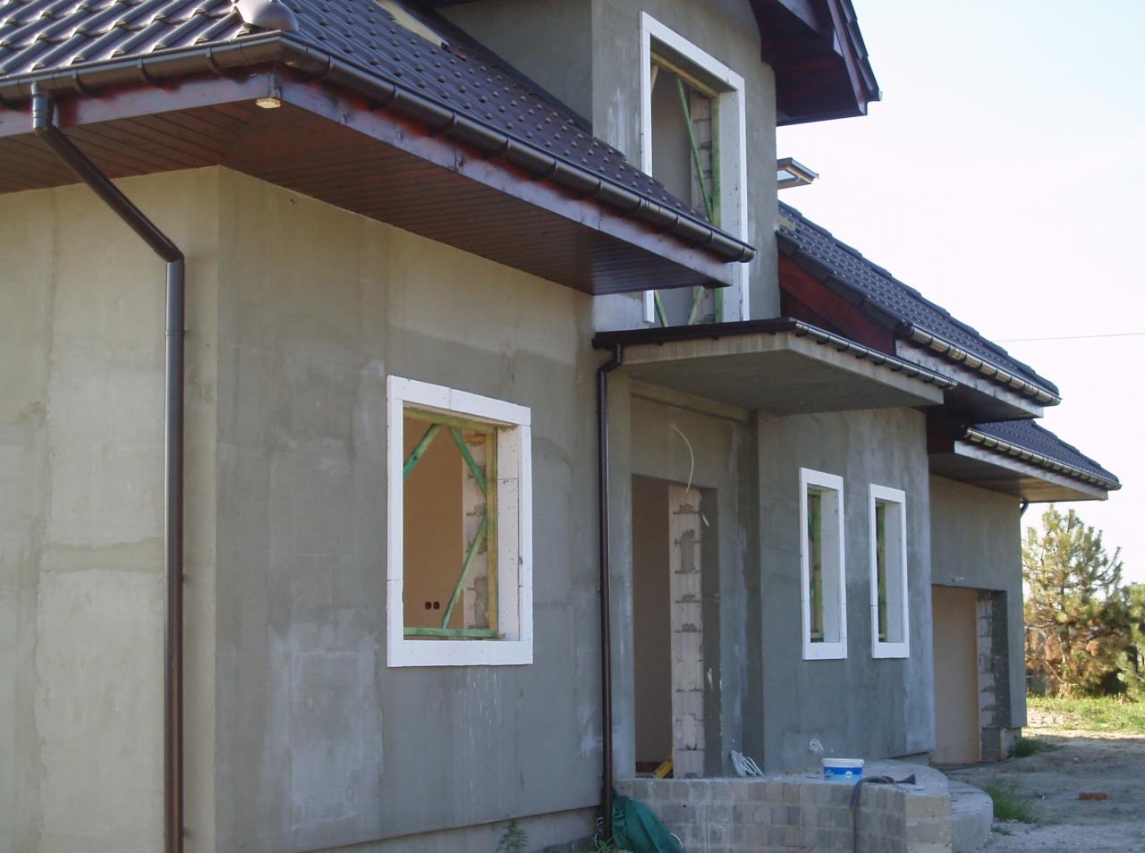 Surowy, dwukondygnacyjny dom z brązową dachówką i oknami w białych ramach przed tynkowaniem, widok z zewnątrz.