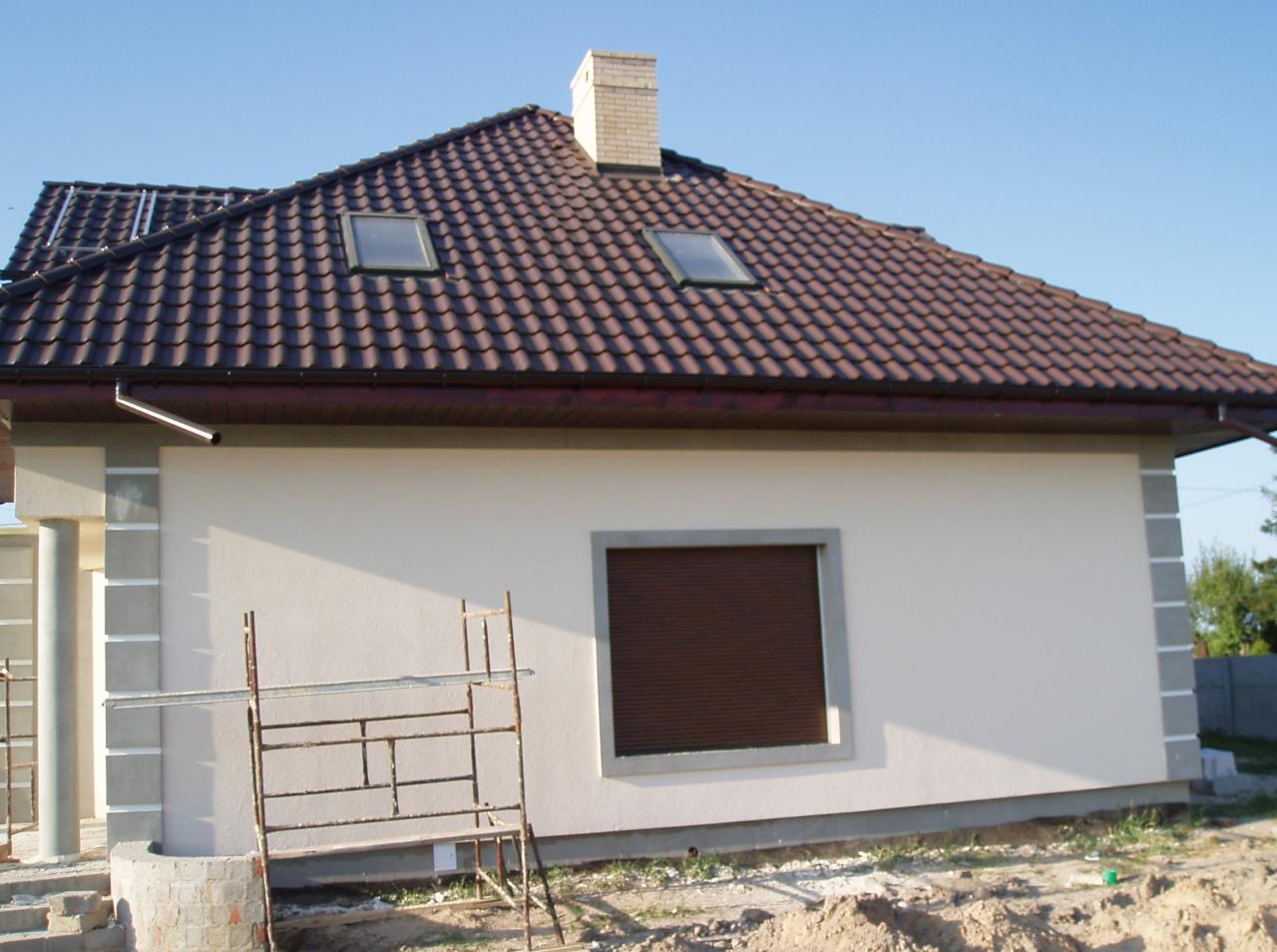Ukończony dom jednorodzinny z brązową dachówką i oknami dachowymi, widok z boku, z fragmentem rusztowania i stertą ziemi na pierwszym planie.