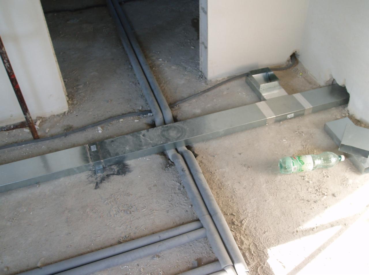 Szare rury i metalowe kanały wentylacyjne ułożone na surowym podłożu w trakcie prac instalacyjnych wewnątrz budynku. Widoczna butelka po napoju.